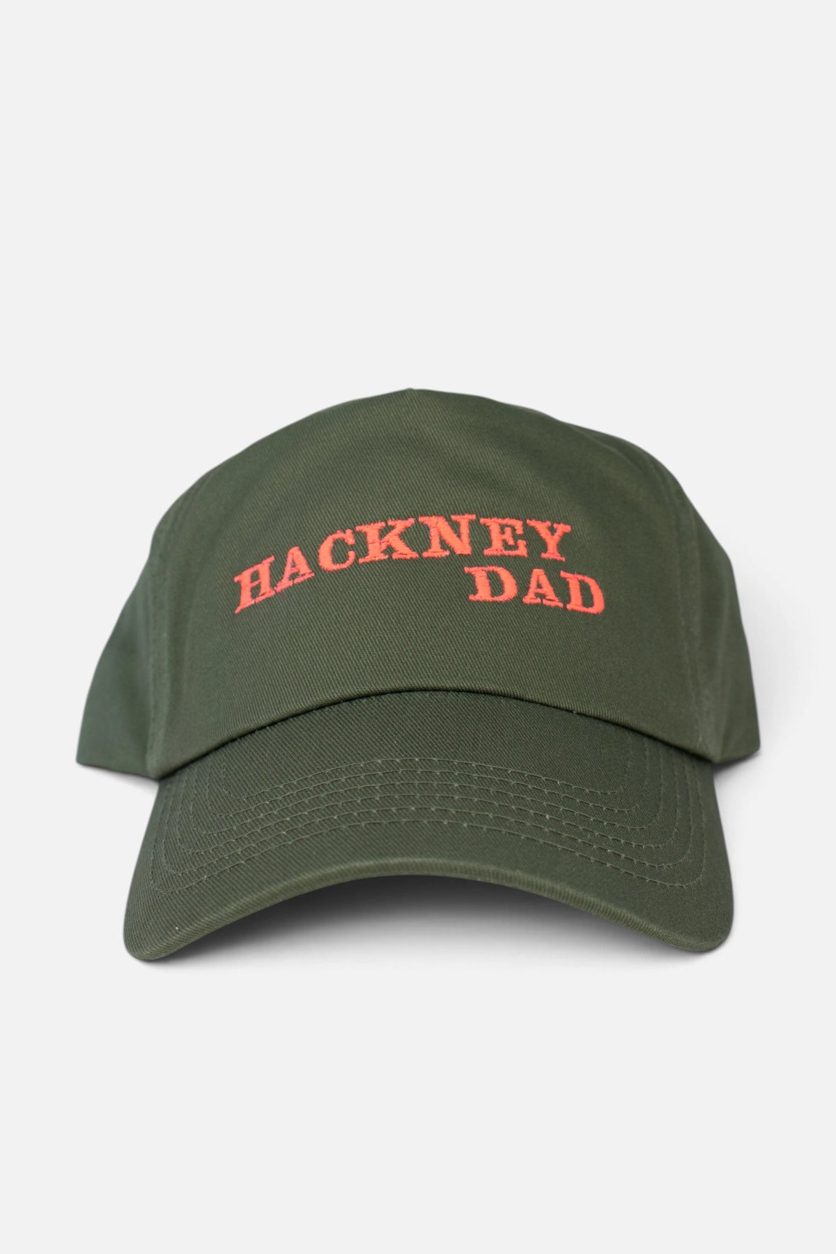 An olive green baseball cap with 'HACKNEY DAD' embroidered in fluorescent coral orange lettering on the front, shown against a light grey background.