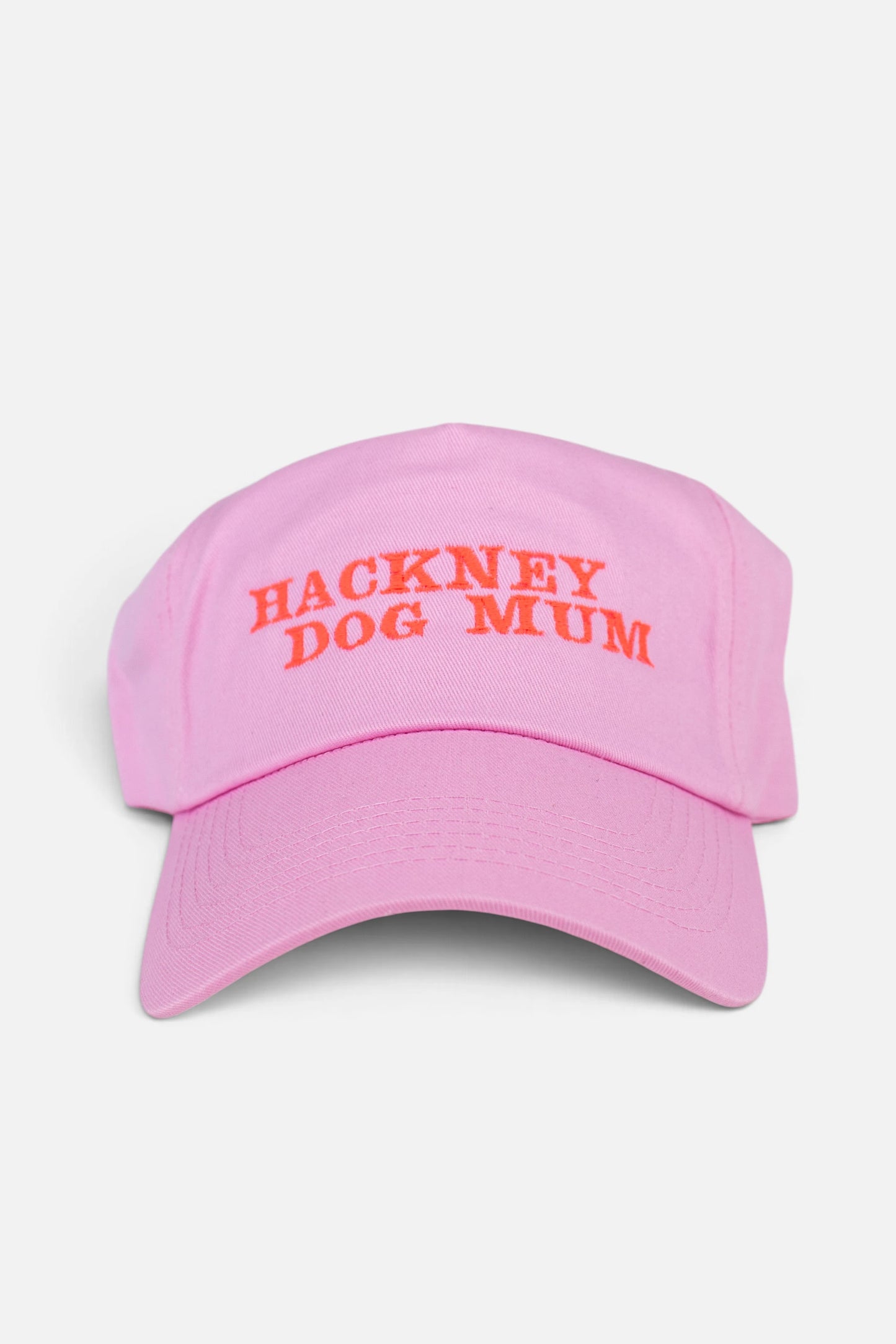 A light pink baseball cap with 'HACKNEY DOG MUM' embroidered in fluorescent coral orange text across the front, shown against a plain light grey background.