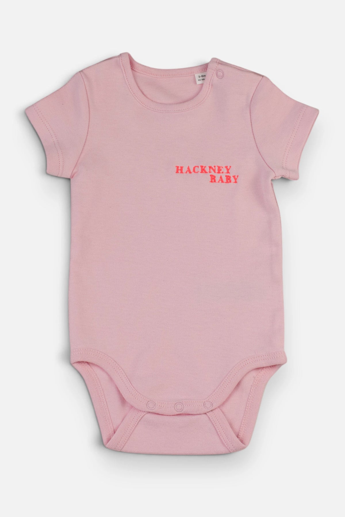 A light pink short-sleeve baby bodysuit with 'HACKNEY BABY' embroidered in fluorescent coral orange on the chest, displayed flat against a light grey background.
