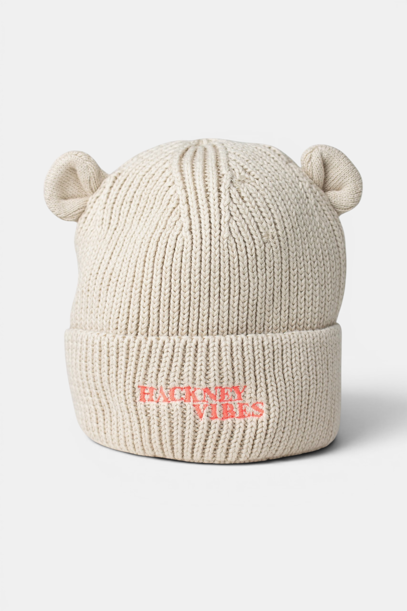 A cream-colored ribbed knit beanie hat with small bear ears on top and the text 'HACKNEY VIBES' embroidered in fluo coral pink on the front cuff, shown against a light gray background.