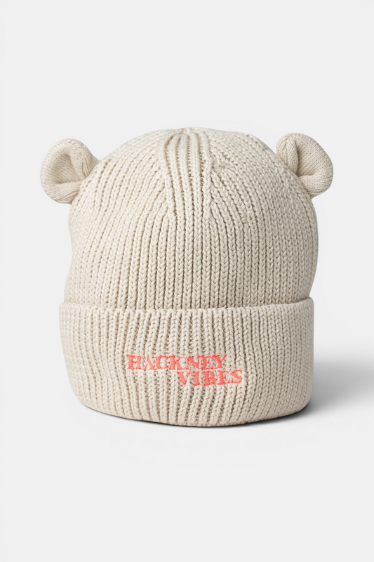A cream-colored ribbed knit beanie hat with small bear ears on top and the text 'HACKNEY VIBES' embroidered in fluo coral pink on the front cuff, shown against a light gray background.