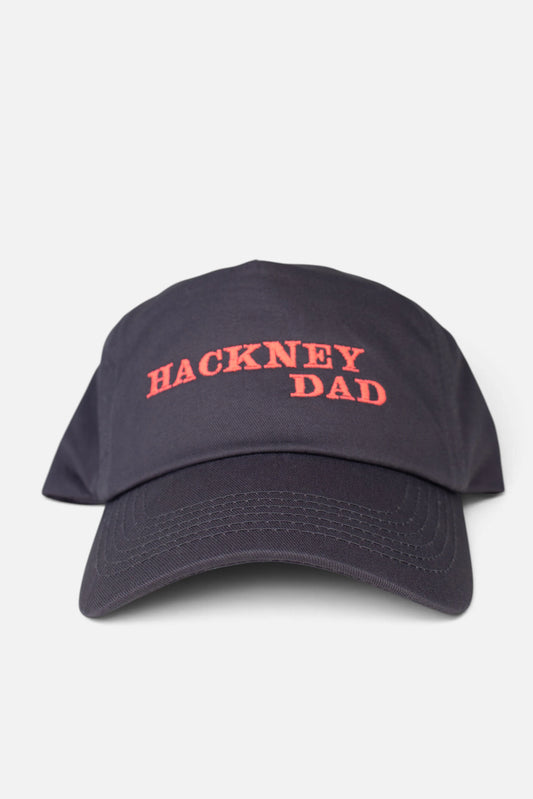 A dark grey baseball cap with 'HACKNEY DAD' embroidered in fluorescent coral orange across the front, shown against a light grey background.