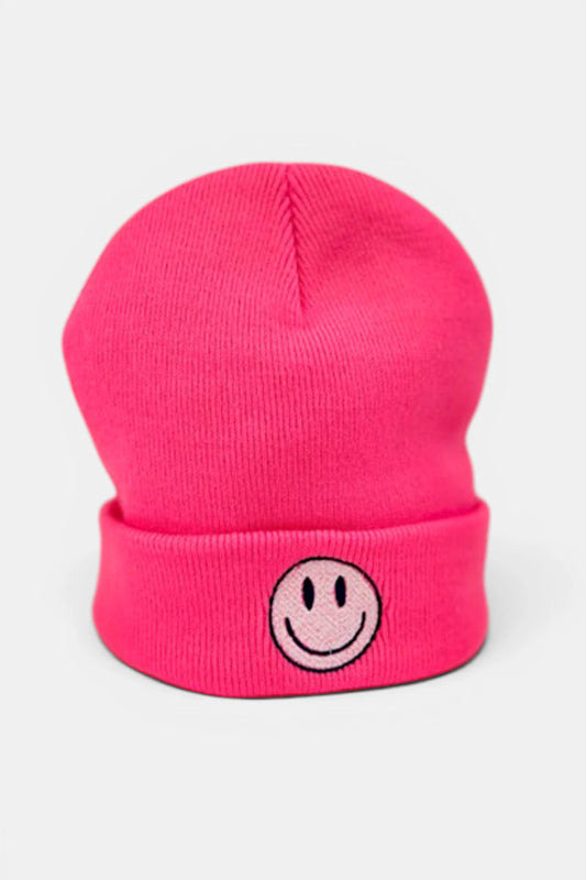 A fluorescent pink beanie hat with a folded cuff featuring a small embroidered smiley face patch in the center, shown against a light gray background.