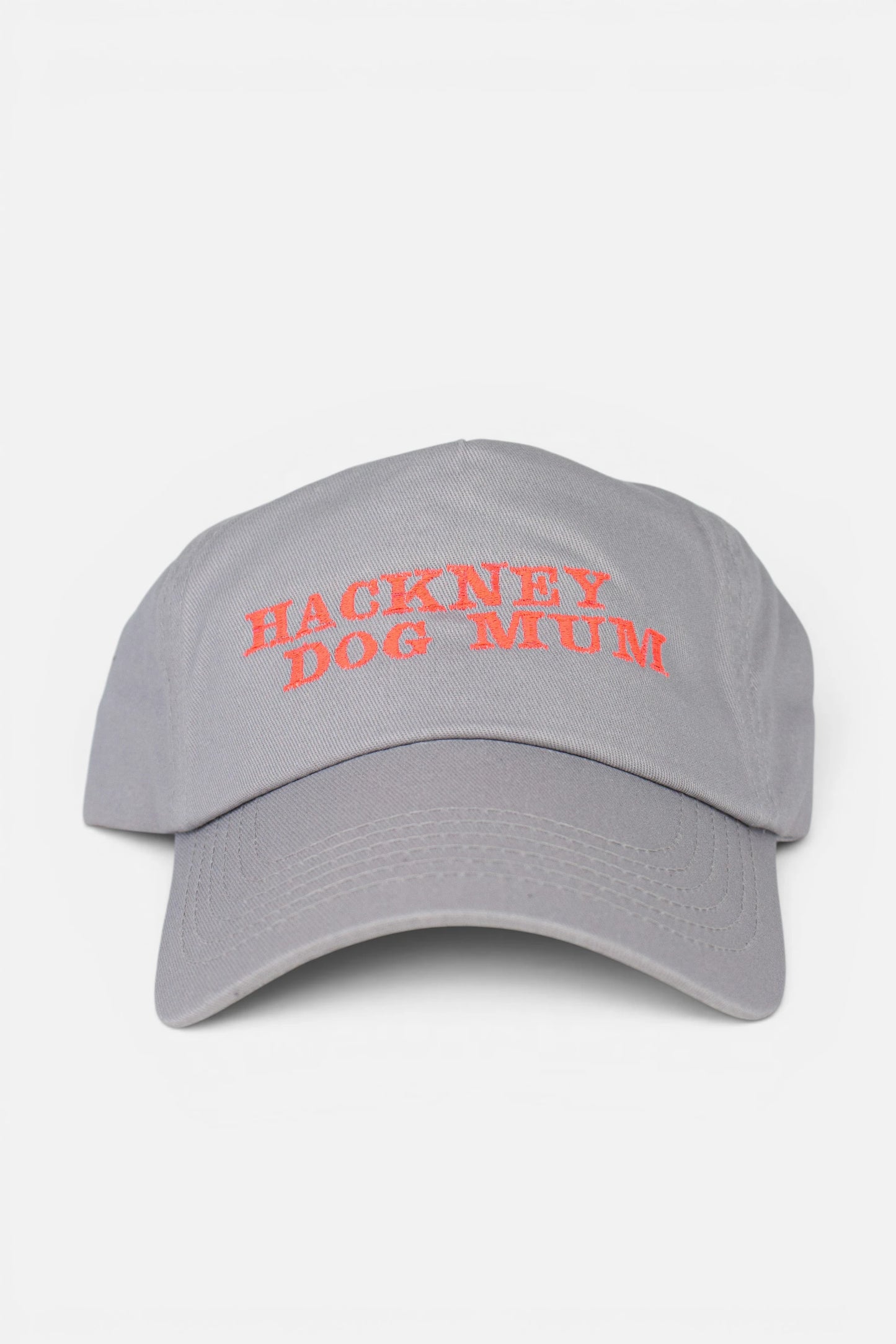A light grey baseball cap with 'HACKNEY DOG MUM' embroidered in fluorescent coral orange text across the front, shown against a plain light grey background. 