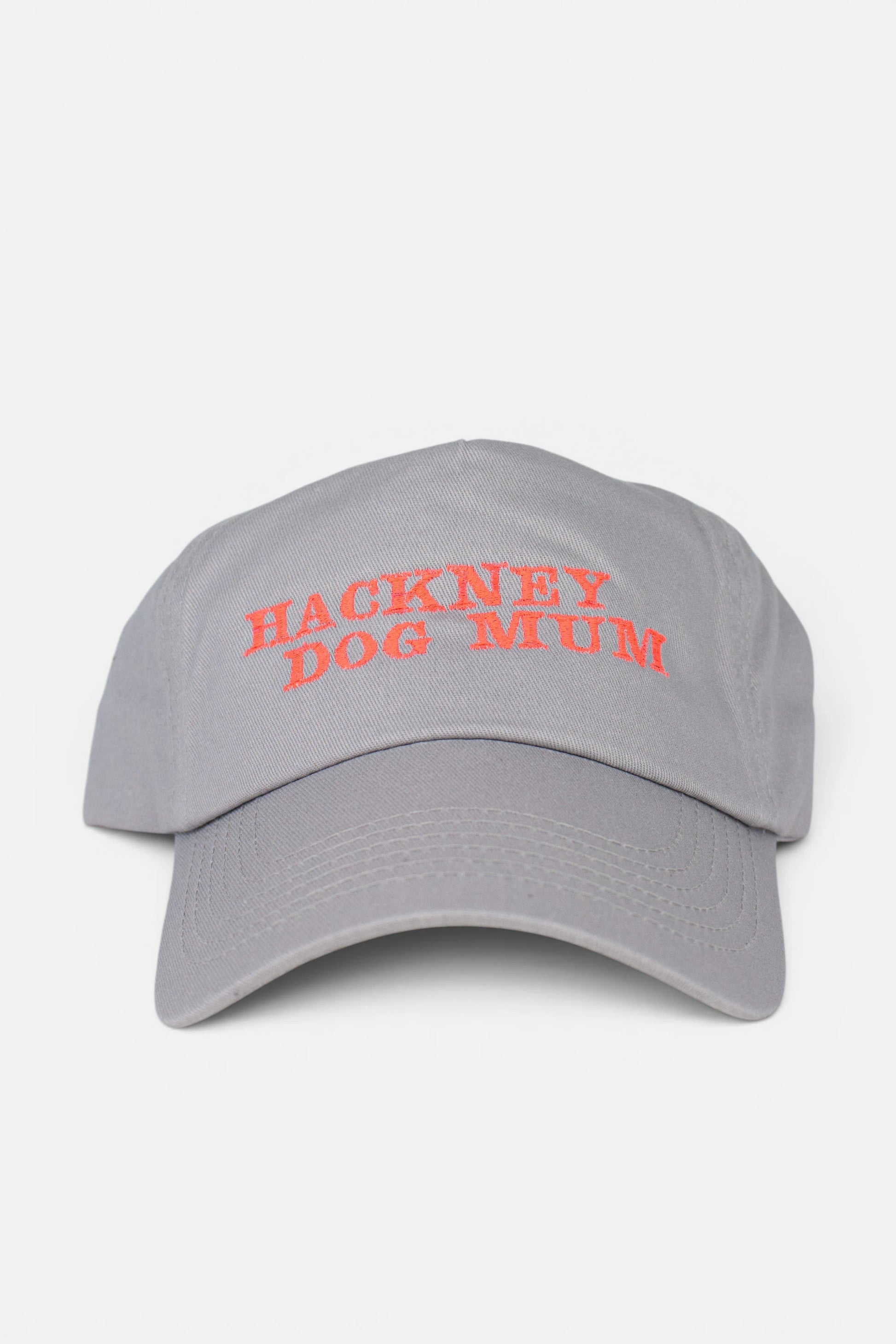 A light grey baseball cap with 'HACKNEY DOG MUM' embroidered in fluorescent coral orange text across the front, shown against a plain light grey background. 