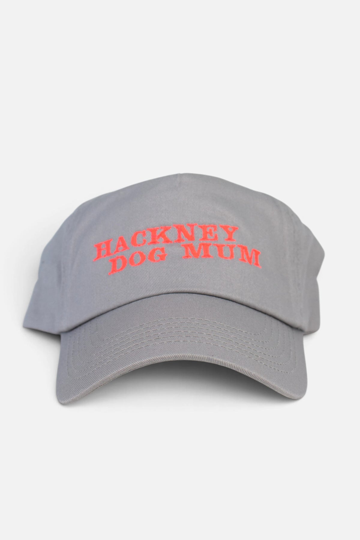 A light grey baseball cap with 'HACKNEY DOG MUM' embroidered in fluorescent coral orange lettering across the front, shown against a plain light grey background.