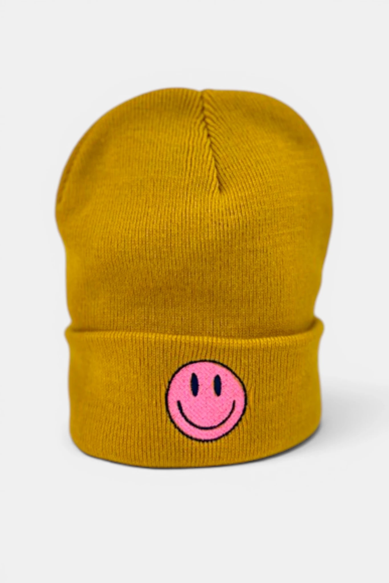 A mustard yellow beanie hat featuring a pink embroidered smiley face on the front cuff, set against a light gray background.