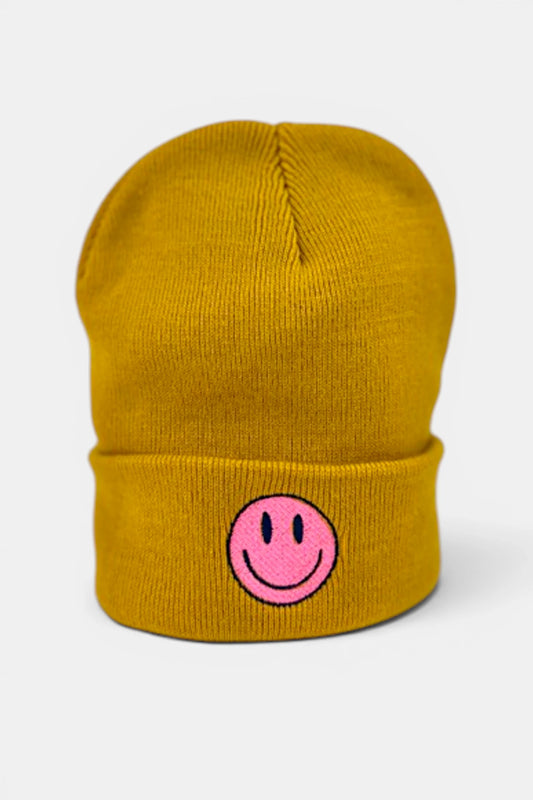 A mustard yellow beanie hat featuring a pink embroidered smiley face on the front cuff, set against a light gray background.