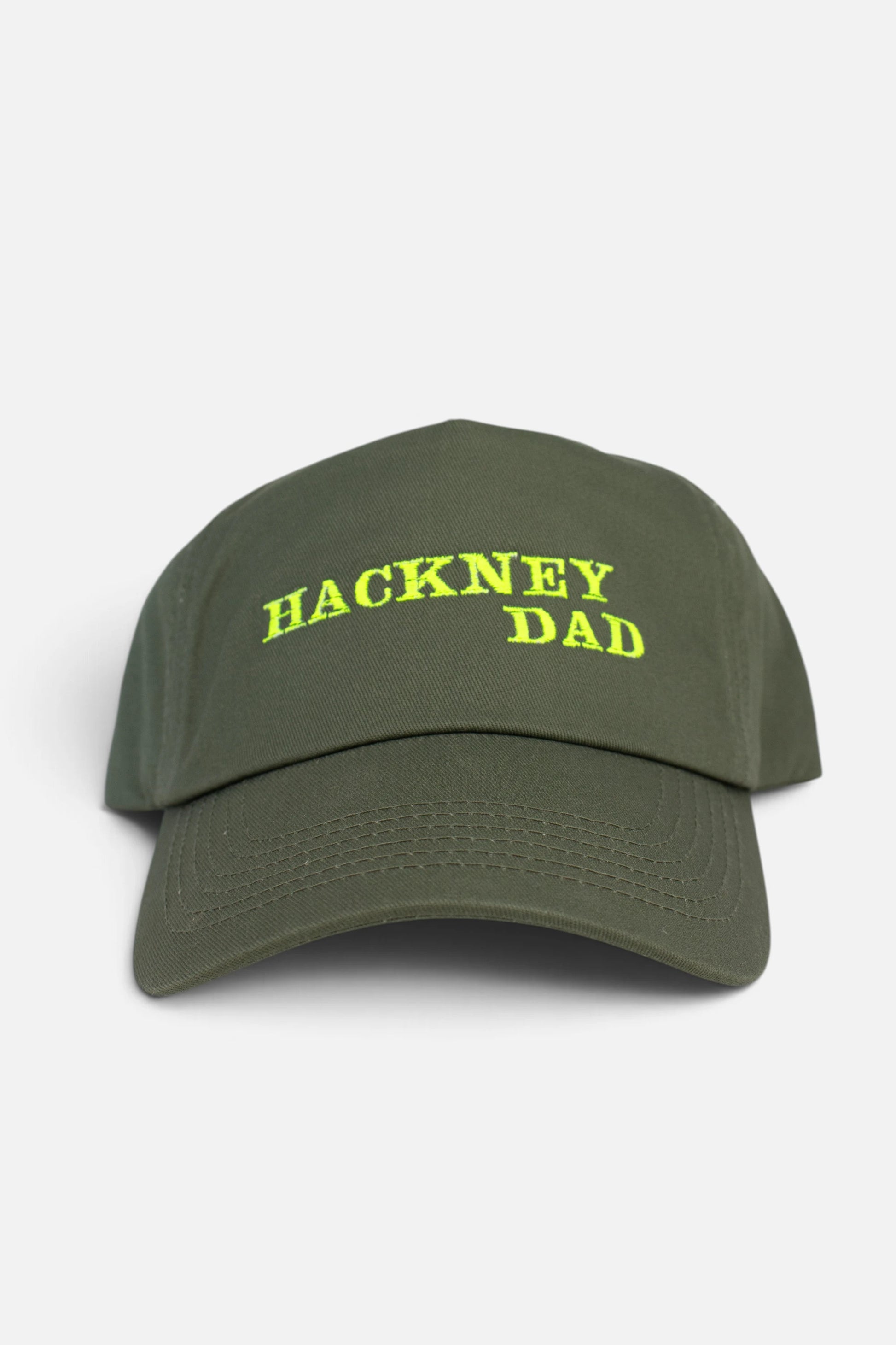 An olive green baseball cap with 'HACKNEY DAD' embroidered in fluorescent yellow text on the front, set against a plain light grey background.