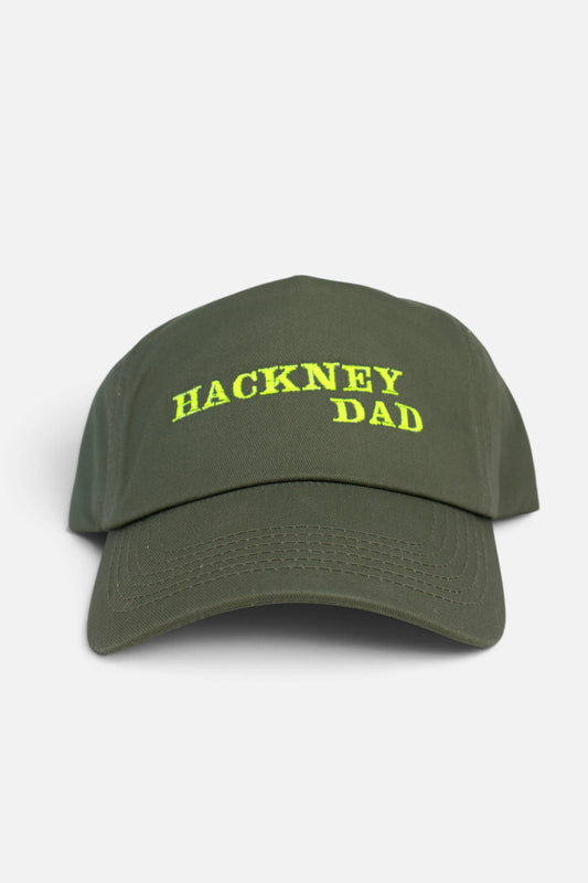 An olive green baseball cap with 'HACKNEY DAD' embroidered in fluorescent yellow text on the front, set against a plain light grey background.