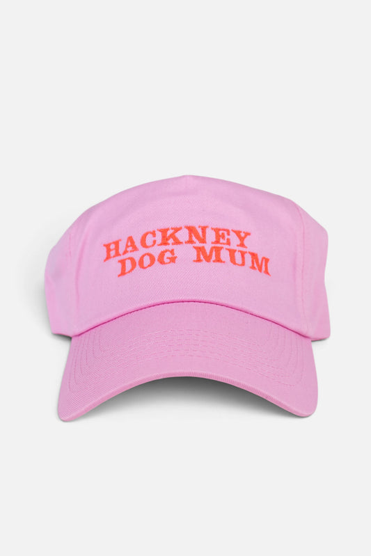 A light pink baseball cap with 'HACKNEY DOG MUM' embroidered in fluorescent  coral orange text across the front, shown against a plain light grey background.