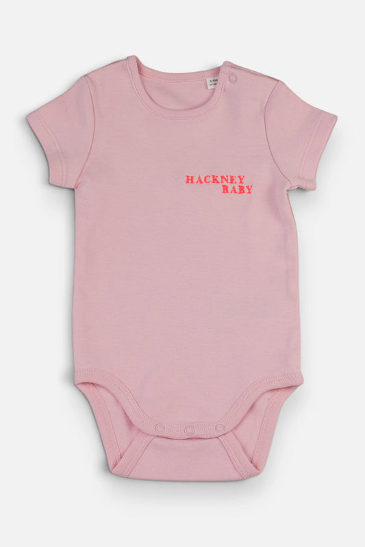 A light pink short-sleeve baby bodysuit with 'HACKNEY BABY' embroidered in fluorescent coral orange on the chest, displayed flat against a light grey background.