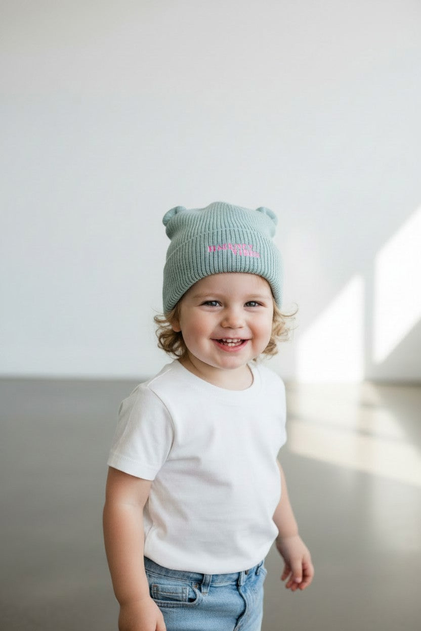 A smiling toddler wearing a light sage green ribbed knit beanie with small ears and pink embroidered text, paired with a white t-shirt and blue jeans, standing in a bright minimalist room.